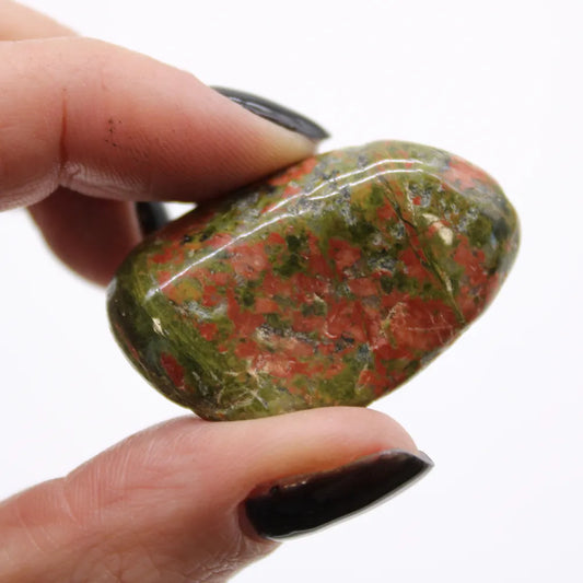 A shiny unakite stone is held between two fingernails with black polish, against a solid white background.