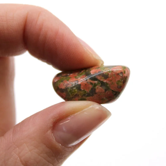 A mottled green and orange unakite stone is being held between a thumb and forefinger against a bright white background.