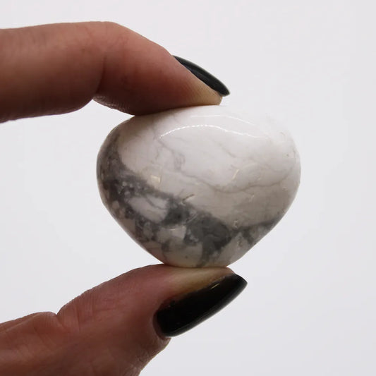 A thumb and forefinger hold a heart-shaped stone in the air. The stone is polished white with grey veins. Background is plain white.