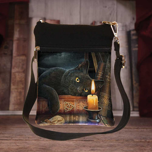 A crossbody bag displays a black cat lounging atop books, near a lit candle. Book titles include “Herbs and Potions” and “The Complete Witch’s Handbook”. The bag is set against a blurred bookshelf background.