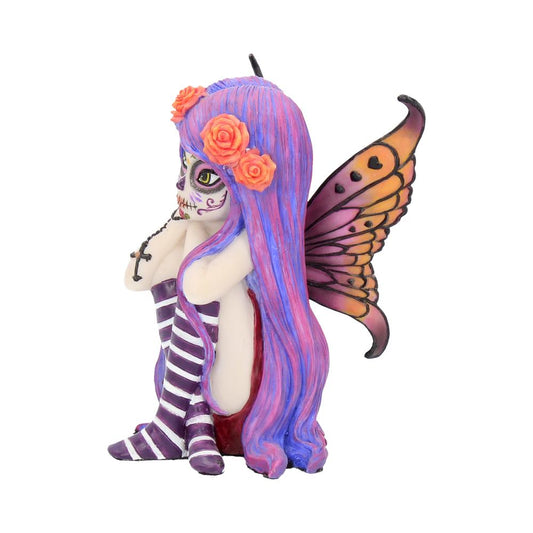 A fairy figurine with Day of the Dead face paint sits holding a black rosary. It has purple and pink hair, orange flowers, striped socks, and orange wings, set against a plain white background.