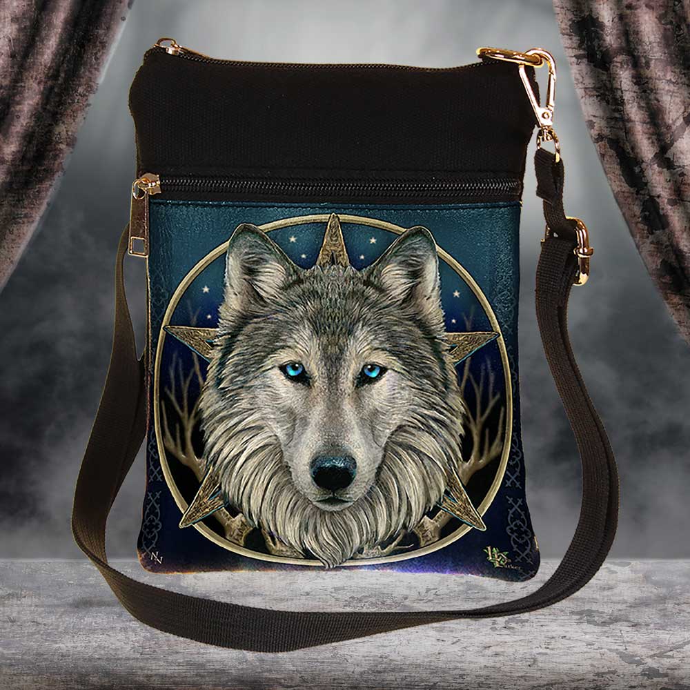 A black crossbody bag displays a wolf’s face in front of a compass star on a blue, star-studded background. The bag features a dark strap and is situated in a neutral setting with draped fabric.