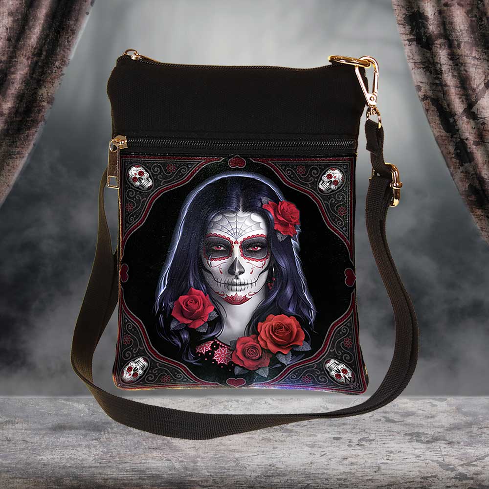 A shoulder bag features a design of a woman with Day of the Dead face paint and red roses. The bag hangs on a rustic surface, with draped fabric in the background.