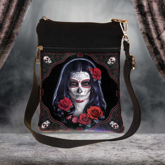 A shoulder bag features a design of a woman with Day of the Dead face paint and red roses. The bag hangs on a rustic surface, with draped fabric in the background.