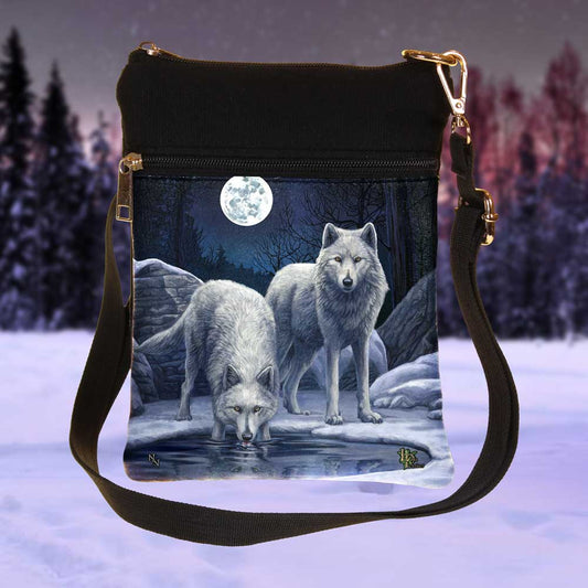 A crossbody bag displays two wolves by water under a full moon and wintery sky. The top of the bag is black, with a zipper closure and shoulder strap.