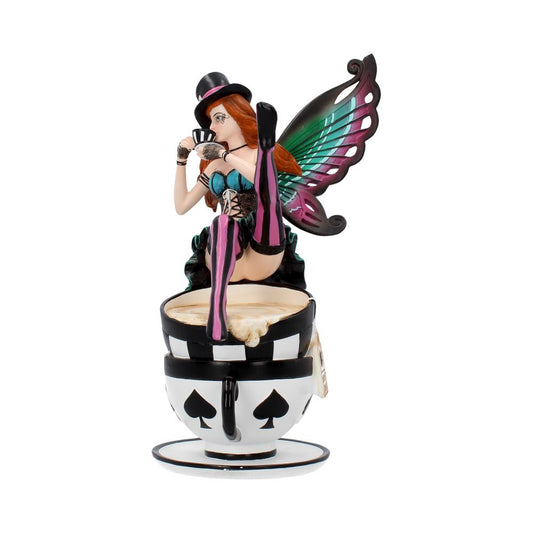 A figurine depicts a fairy with colorful wings sitting on a large, patterned teacup. She wears a top hat and corset, holding playing cards in a whimsical setting.