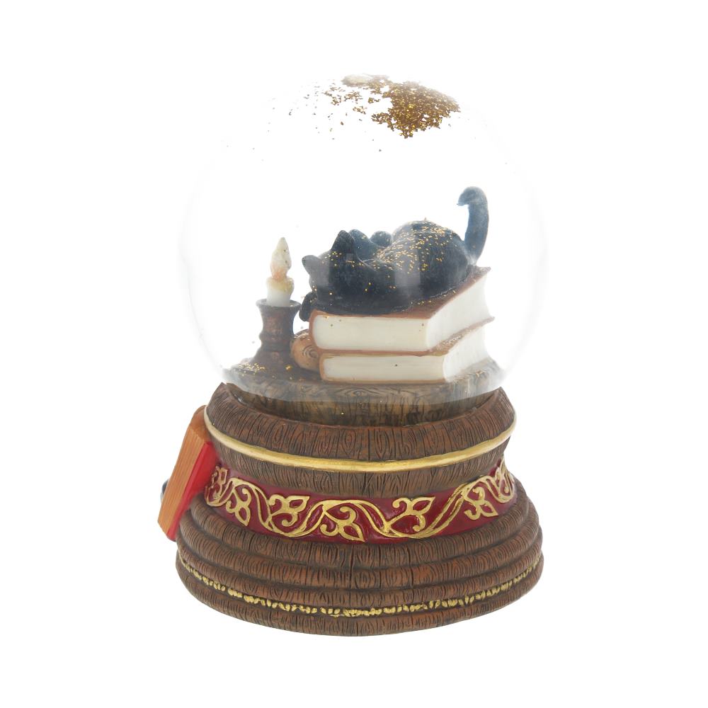 A snow globe contains a black cat, lounging on top of stacked books next to a candle. Gold glitter floats inside, and the base is decorated with a patterned red ribbon.