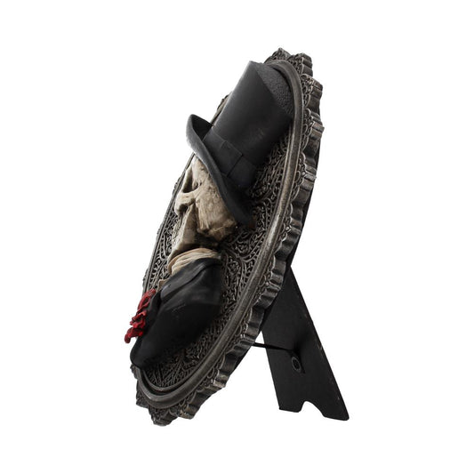 A skull wearing a top hat is affixed to an ornate, circular plaque; it stands on a small, black stand.