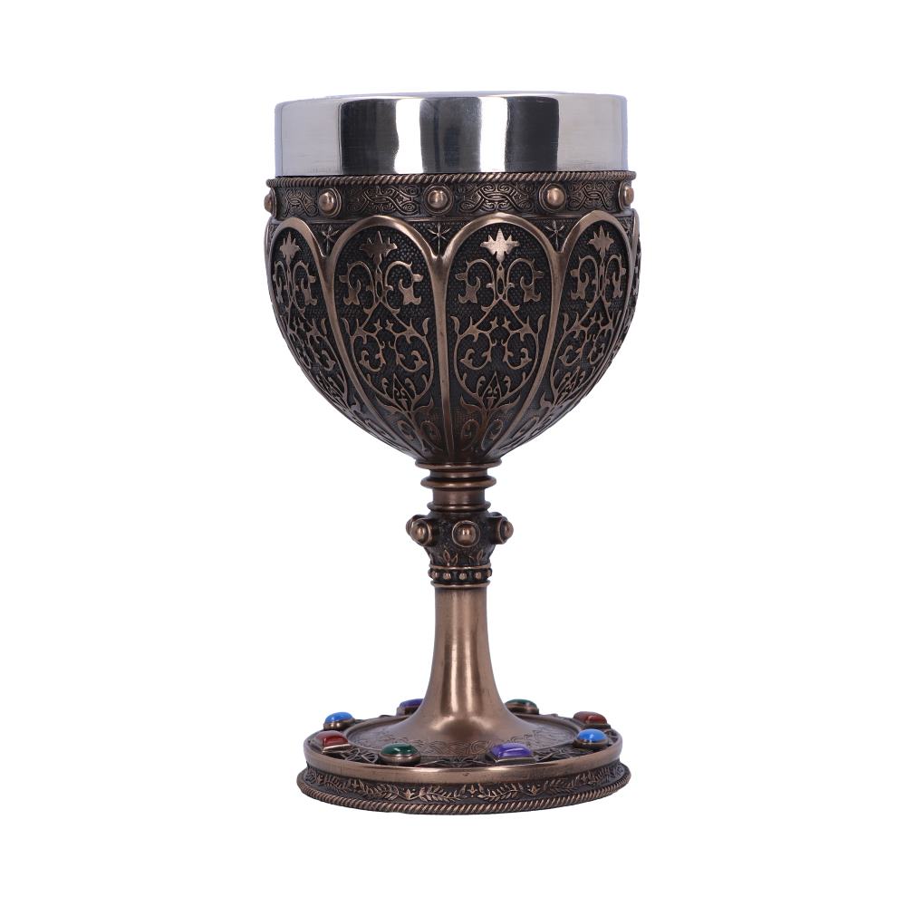 An ornate, stemmed chalice stands on a white surface. The cup features a silver-colored top, bronze-colored carvings, and the base is decorated with colorful gems.