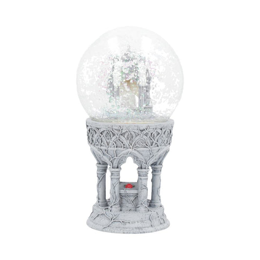 A snow globe containing a miniature castle sits atop an ornate, grey, gothic-style pedestal. A small red object rests beneath the globe's base.
