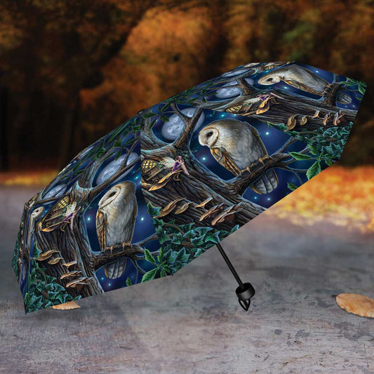 A compact umbrella, featuring owls perched on a tree at night, rests on a paved surface against a blurred autumnal background.