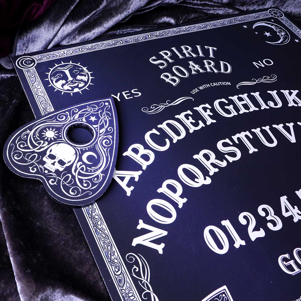 A spirit board sits on a velvet cloth, featuring a detailed planchette with a skull design. The board displays letters, numbers, and text: "SPIRIT BOARD, USE WITH CAUTION, YES, NO, GOODBYE."