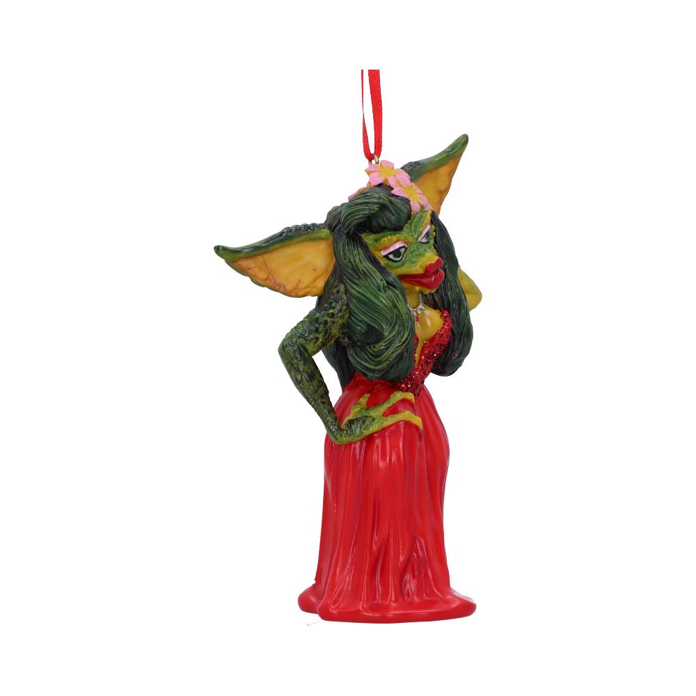 A green creature ornament with long hair wears a red dress and poses confidently, hanging from a red ribbon. It's decorated with flowers and stands against a plain white background.