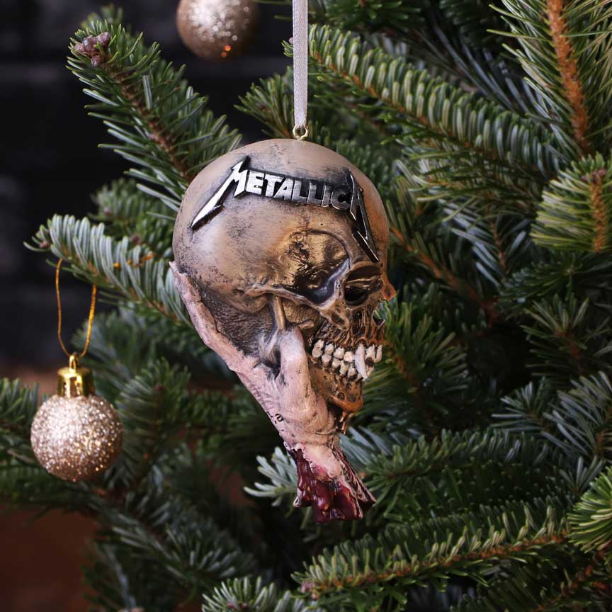 Skull-shaped Christmas ornament labeled "Metallica" hangs from a ribbon on a fir branch, showing exposed teeth and a bloody neck stump beside a glittery gold bauble