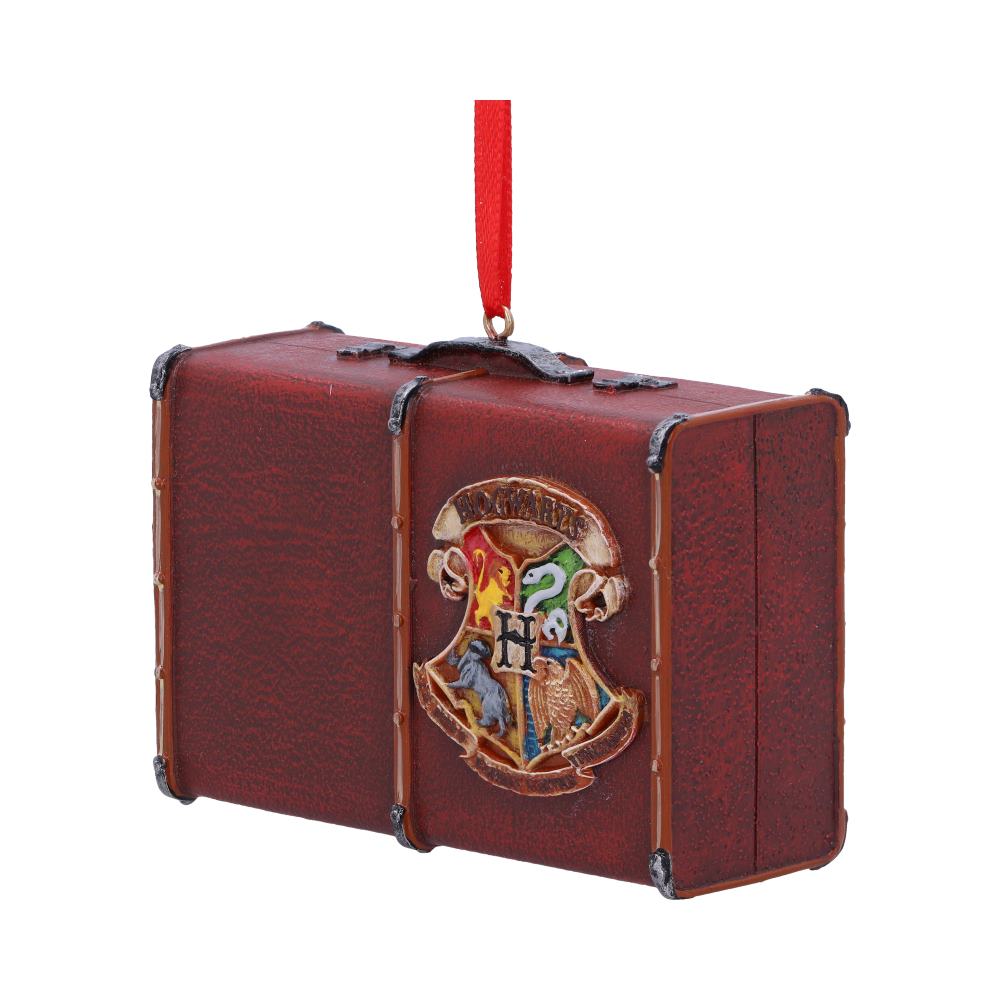 A small brown suitcase ornament hangs from a red ribbon, featuring a colorful crest with an "H" and animals symbolizing four houses, set against a plain white background.