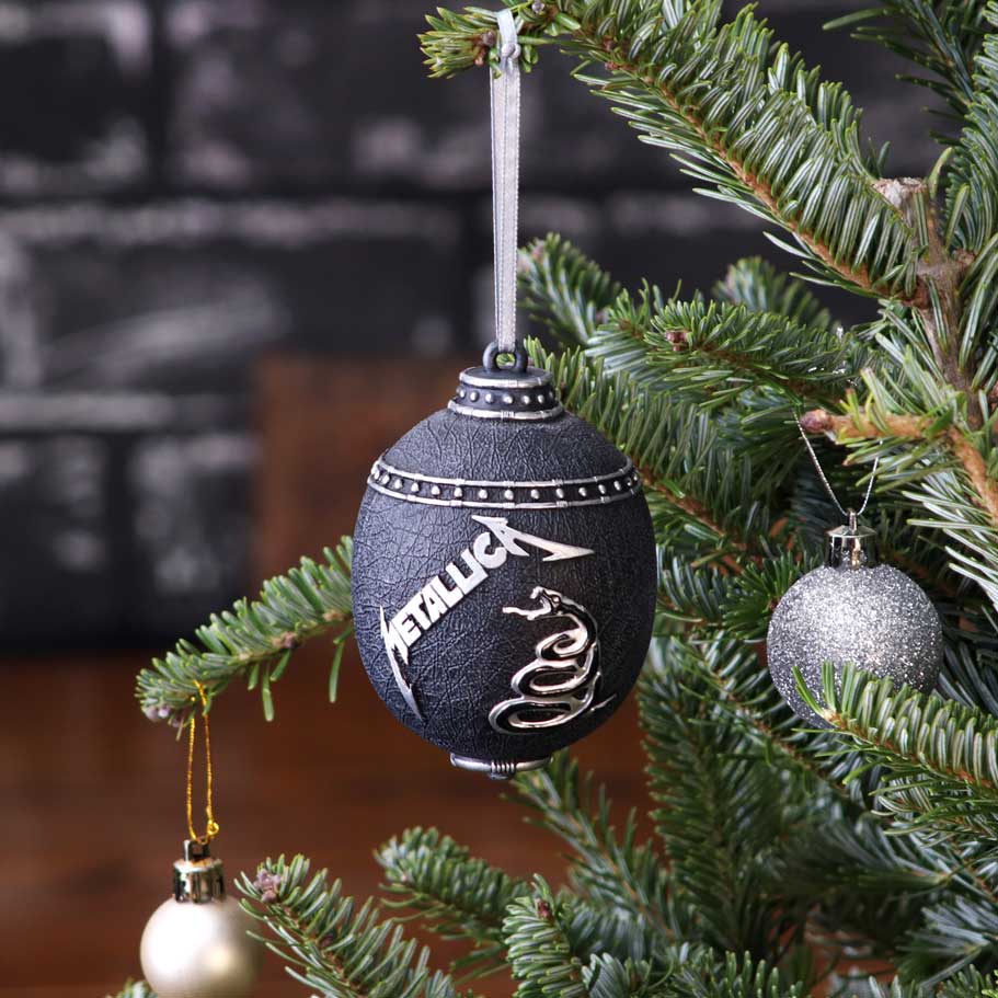 Black spherical ornament labeled "METALLICA" hangs from a silver ribbon on a fir branch, surrounded by glittery silver and pearl baubles against a blurred dark brick and wooden background.