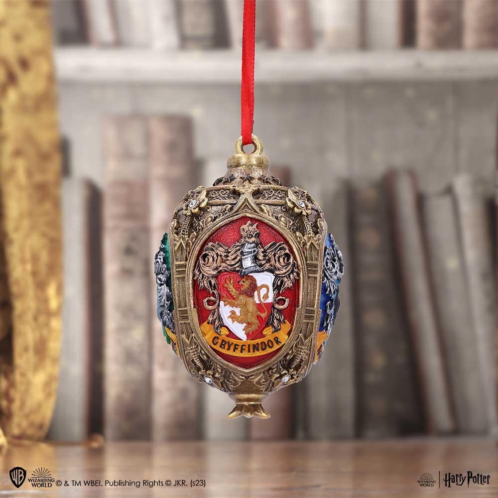 An ornate Gryffindor-themed ornament hangs from a red ribbon, featuring a lion crest and surrounded by intricate designs. It is set against a blurred background of books on a shelf. Text: "GRYFFINDOR", and credits to "™ & © WBEI. Publishing Rights © JKR.".