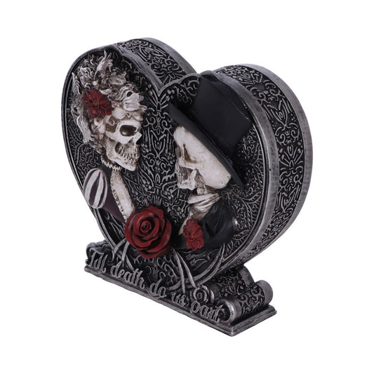 A heart-shaped figurine depicts two skeletons embracing; one wears a top hat. The base reads, “Till death do us part.”