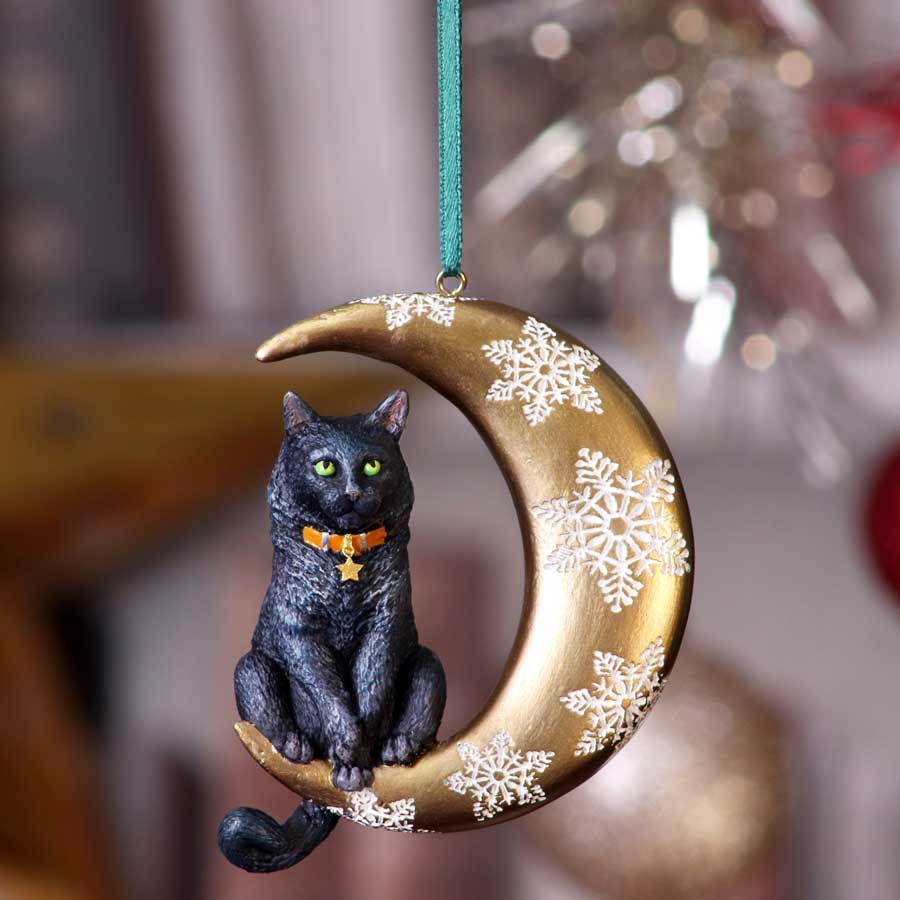 Black cat ornament with green eyes sits on a gold crescent moon decorated with white snowflakes, hanging from a teal ribbon against a softly blurred holiday-lit indoor background.
