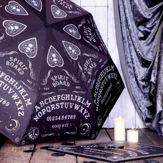 A black umbrella, patterned with a repeating ouija board design, sits closed near lit candles and a smaller ouija board; all on a wooden floor with a velvet backdrop.