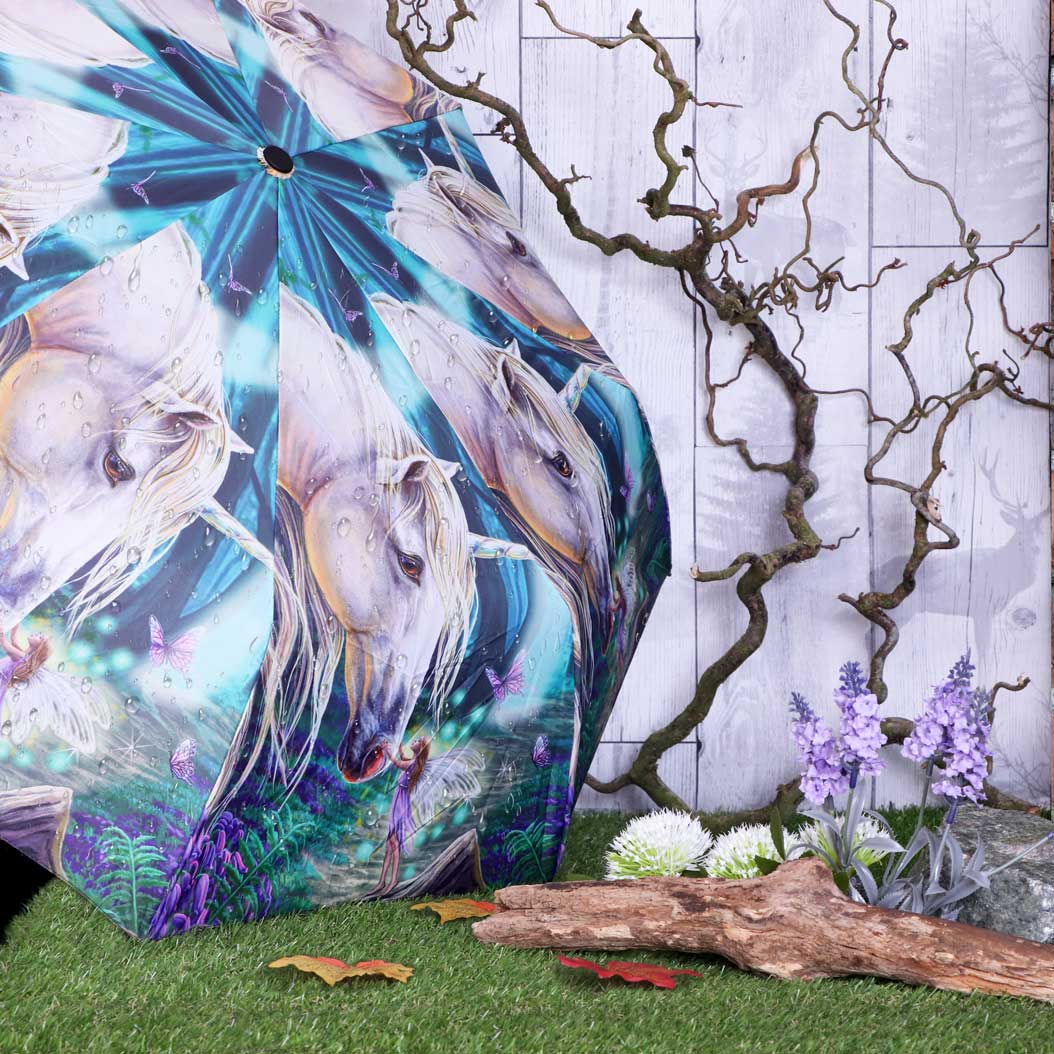 Umbrella decorated with white unicorns and butterflies against a fantasy landscape, leaning on artificial grass. Nearby are a twisted branch, purple flowers, and a wooden plank, set against a pale wooden backdrop.