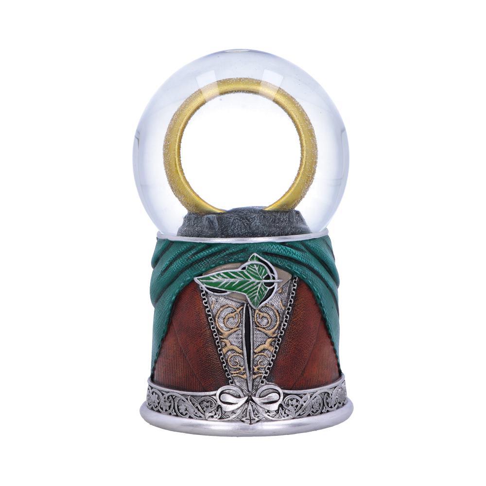 A clear glass globe encases a golden ring, set on a base resembling fabric and leather with decorative silver elements, including a green leaf emblem. The background is plain white.