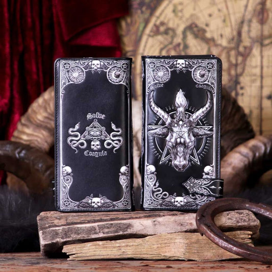 Two long black wallets, displaying occult imagery, rest on a wooden surface near a horseshoe. One features snakes and an eye; the other, a goat's head. The words "Solve" and "Coagula" appear on one wallet.