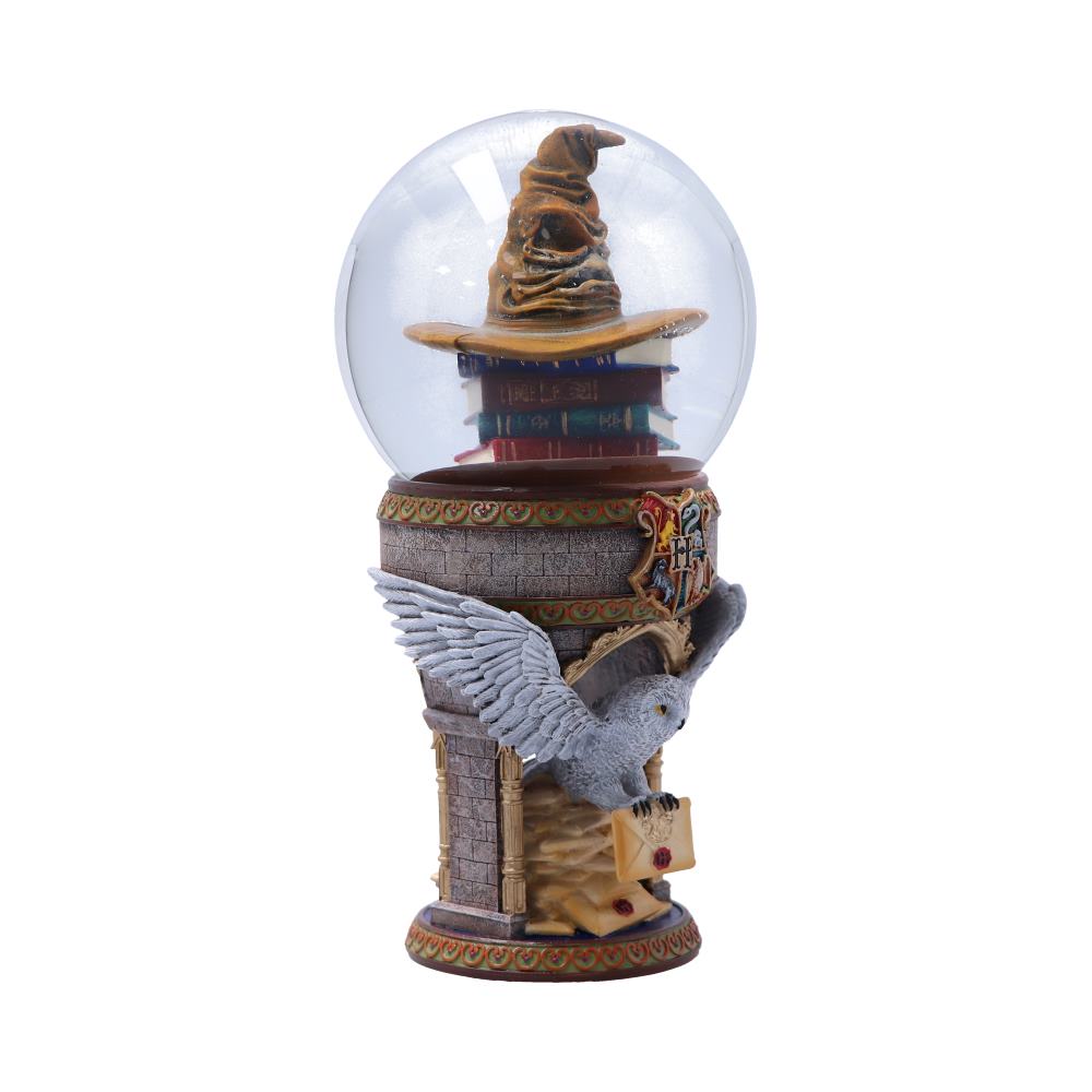 A snow globe contains a sorting hat atop stacked books. The globe rests on a detailed castle base with a flying owl holding a letter. Decor includes a "Hogwarts" crest.