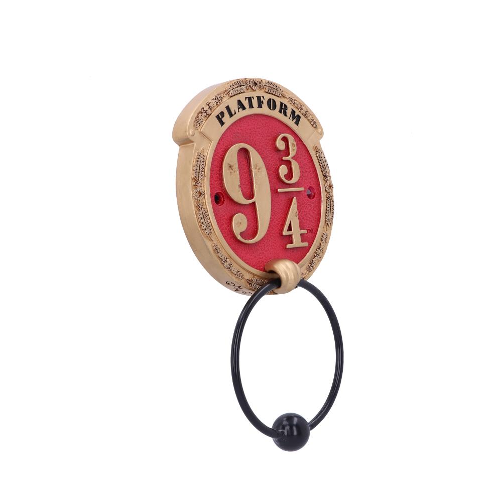 A decorative wall hook features a circular plaque labeled "PLATFORM 9 3/4" in golden letters on a red background, with an attached black ring below, isolated against a plain backdrop.