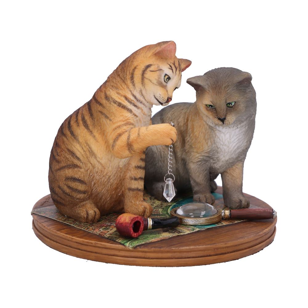 Two cat figurines are seated on a circular base with map-like details. One cat holds a crystal on a chain, surrounded by a pipe and a magnifying glass.