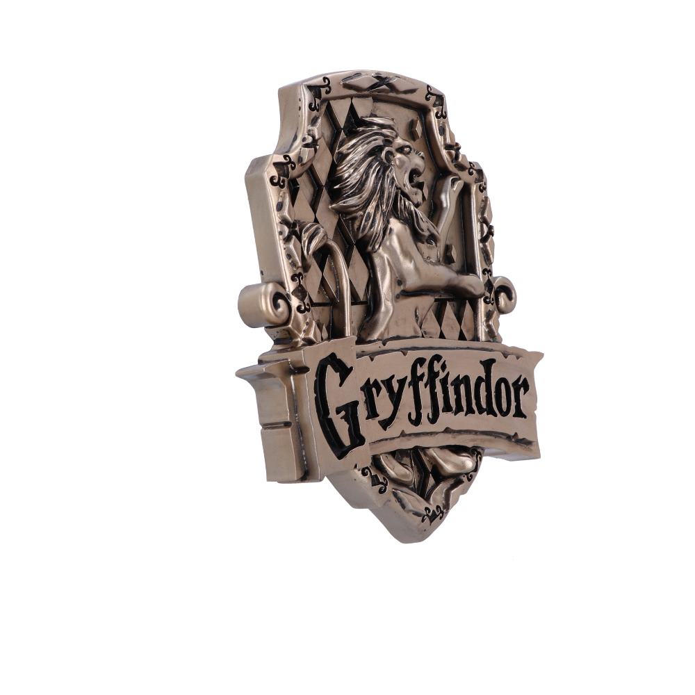 A bronze-colored emblem features a lion in mid-roar with the word "Gryffindor" inscribed in bold black letters on a banner, set against a decorative, ornate background.