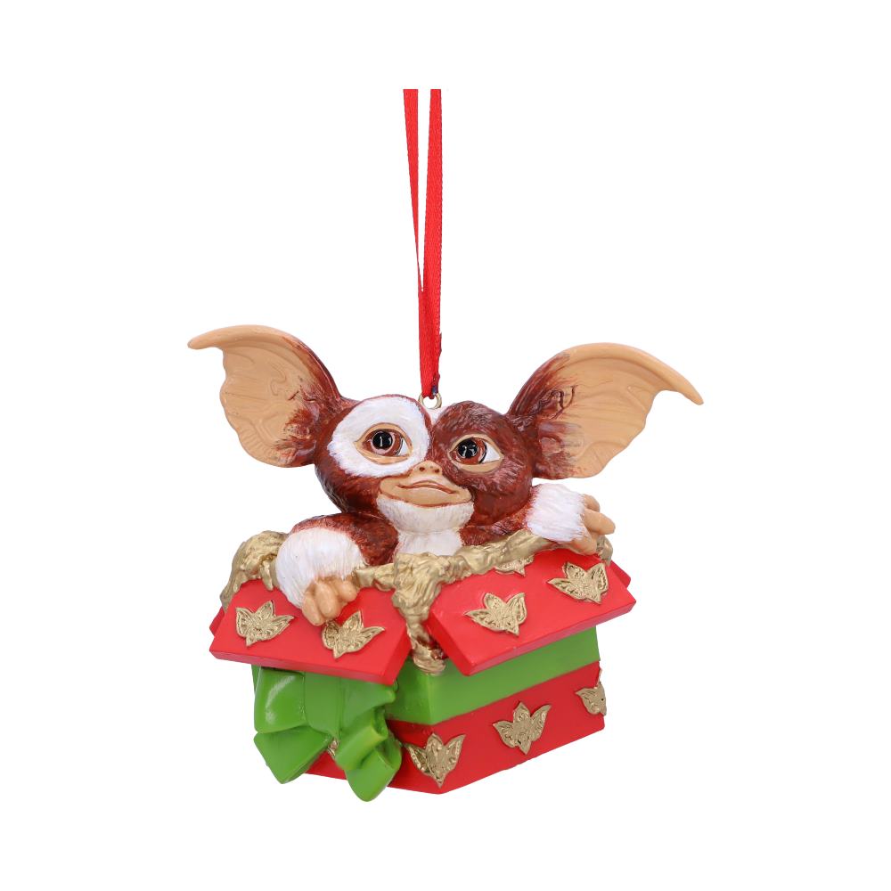 A small creature with large ears peeks out of a decorated red and green gift box ornament, adorned with golden butterflies, hanging from a red ribbon.