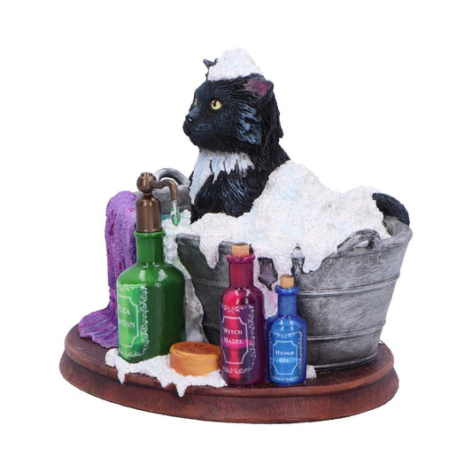 A black cat figurine is enjoying a bath in a small metal tub, surrounded by soap, a towel, and bottles labeled "TEA POTION," "WITCH HAZEL," and "HYSSOP OIL".