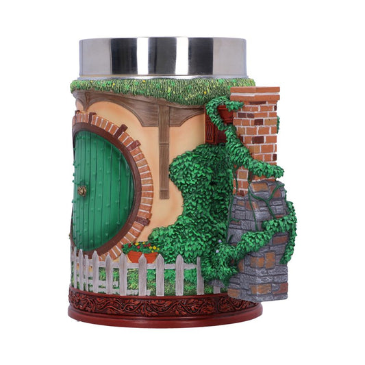 A cylindrical container with a polished metal lid features a decorative façade resembling a hobbit house, including a green door, brick chimney, lush greenery, and a small white picket fence.