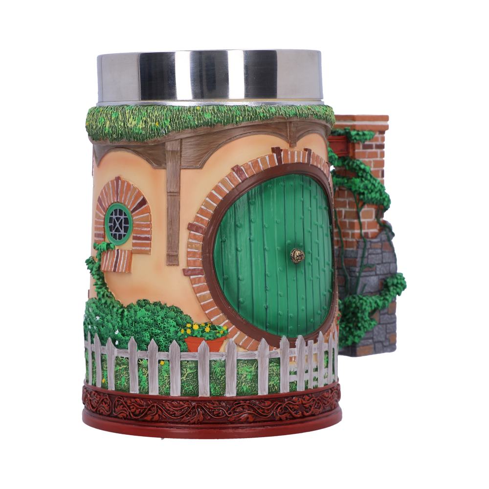 A mug shaped like a hobbit house, featuring a green round door and small window, surrounded by greenery and a picket fence. The top is metallic.