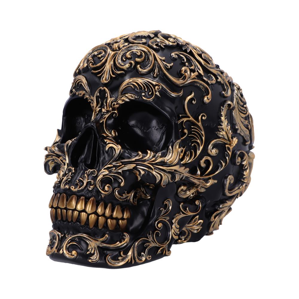A black skull is adorned with intricate, swirling gold patterns covering its surface, set against a plain white background. The design features elaborate floral motifs and gold teeth.