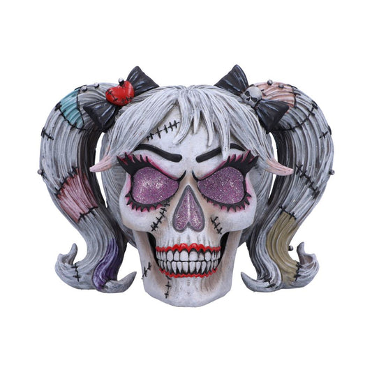 A sculpted skull, styled as a girl with pigtails, displays glittering eyes. The skull features stitched details and rests against a white background.