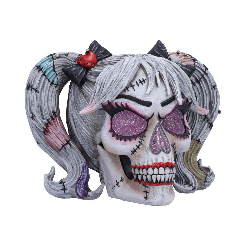 A sculpted skull, styled as a girl with pigtails, sits against a white background. The skull has stitched details, sparkly eyes, and a painted red mouth.