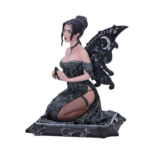 A dark-haired fairy figurine, kneeling, holds an object; it has ouija-board-patterned wings. The figurine rests on a dark, ornate base. The wing displays "NO" and part of the alphabet.