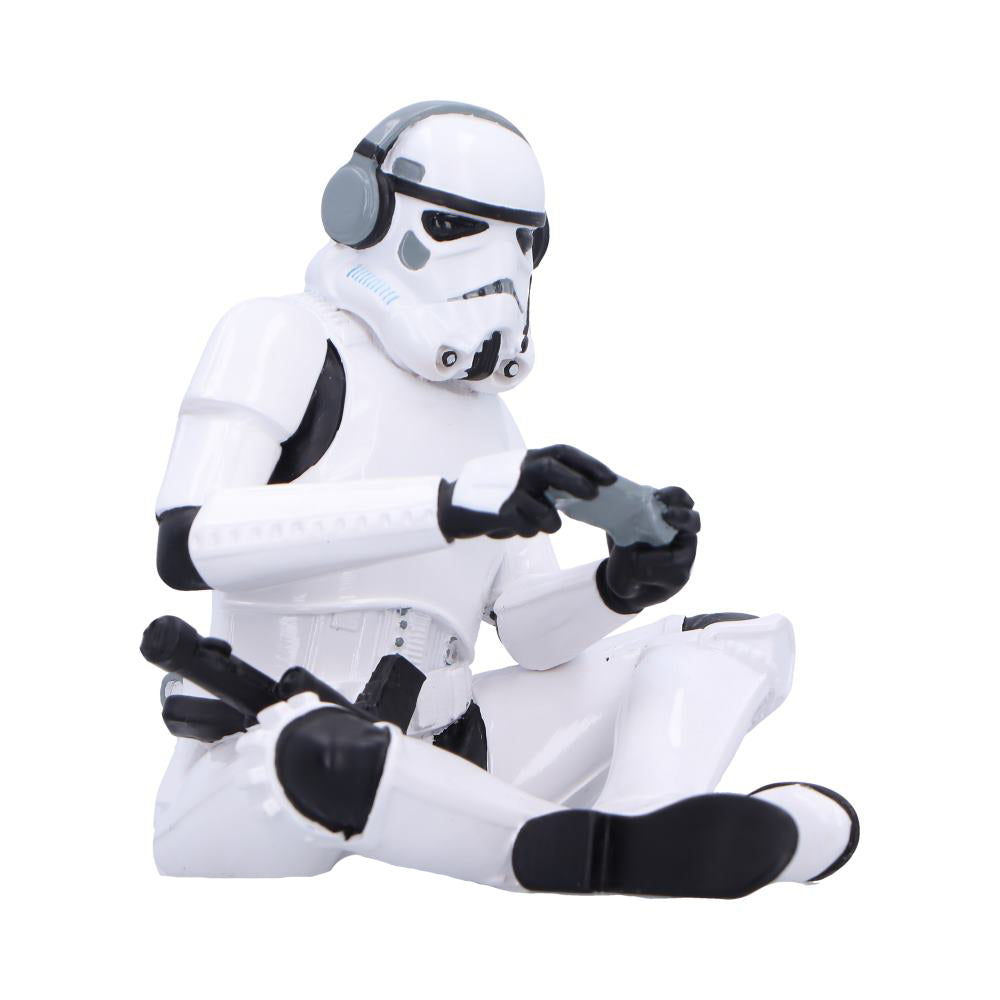 A white-armored Stormtrooper toy sits cross-legged wearing gray headphones and holding a small game controller as if playing, photographed against a plain white background.