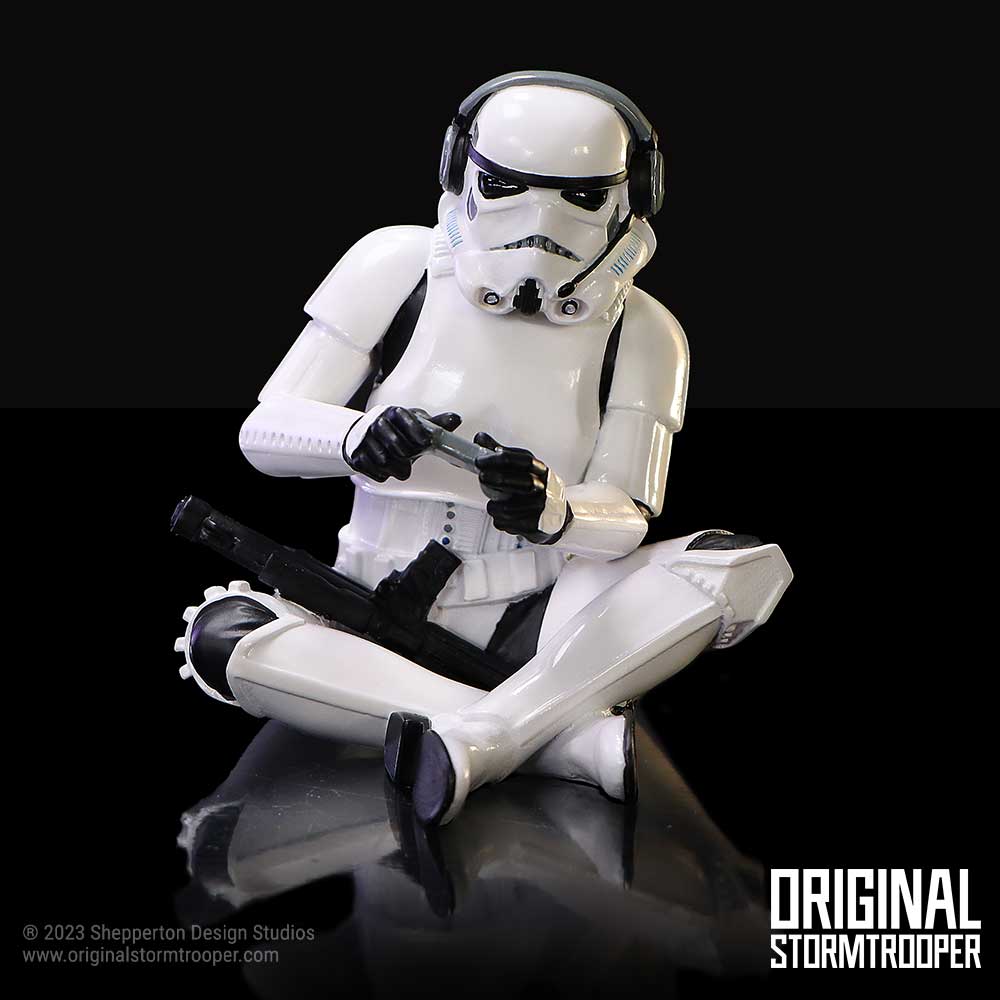 White stormtrooper figurine sits cross-legged, holding and inspecting a small device on a reflective black surface with a black background, text: © 2023 Shepperton Design Studios www.originalstormtrooper.com ORIGINAL STORMTROOPER