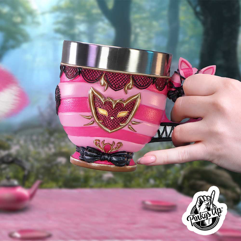 A pink striped decorative teacup with cat-mask emblem is held by a hand over a woodland table; text on image reads "Pinky's Up™".