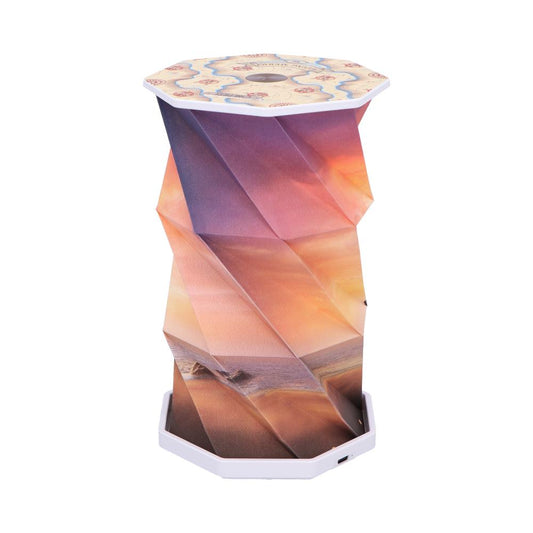 Octagonal portable folding lamp unfolds into a twisted origami column, displaying a printed sunset beach scene on pleated panels, capped by white octagonal top and base with USB charging port.