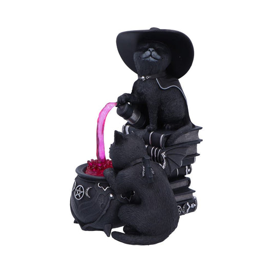 Black cat figurine wearing a witch hat pours glowing pink liquid from a vial into a cauldron while another cat watches, all positioned atop stacked spell books on white background.