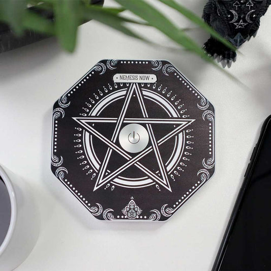 A black octagonal device printed with white pentagram and central power symbol labeled "NEMESIS NOW" sits on a white desk beside a mug and smartphone, partially obscured by plant leaves.
