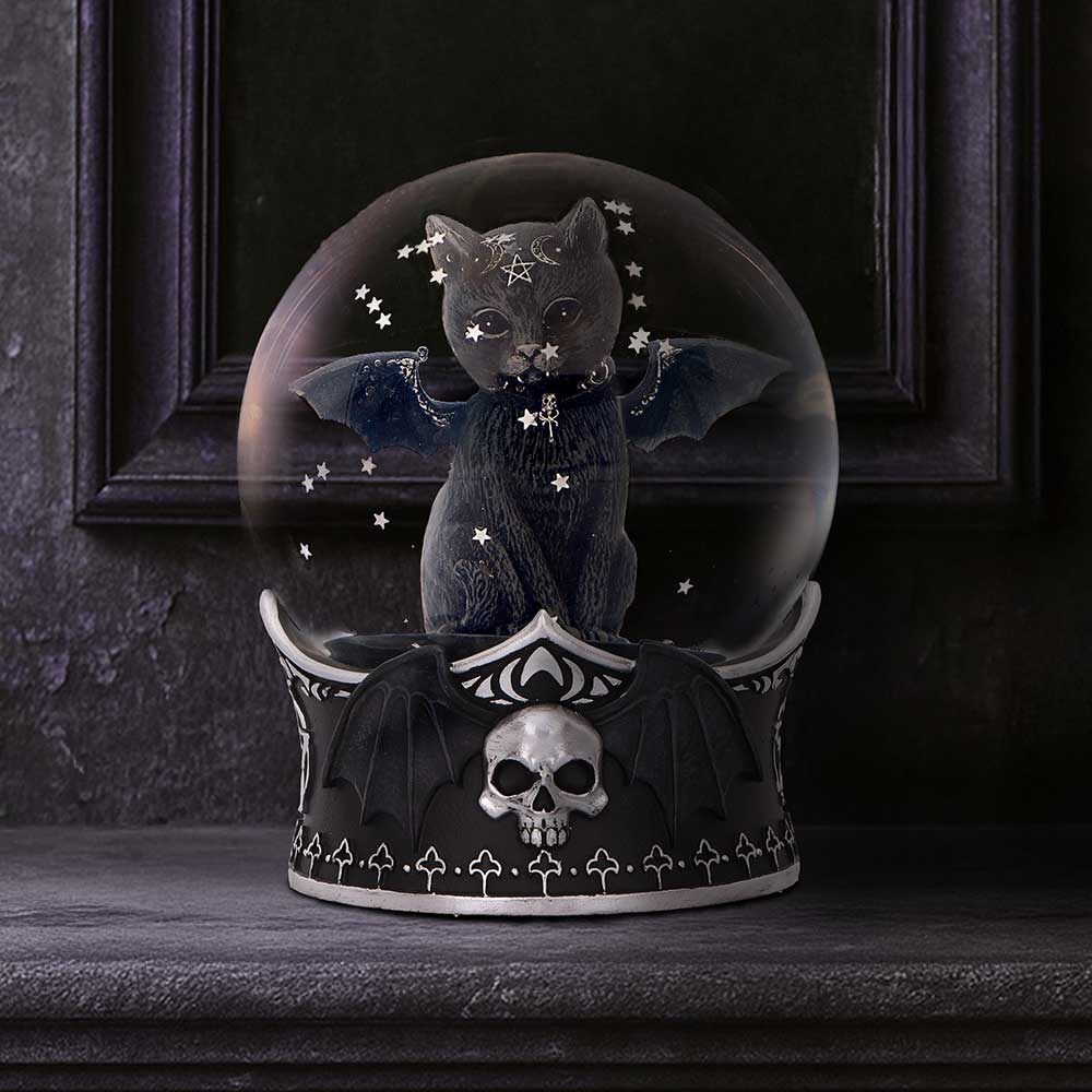 A black cat figurine with outstretched bat wings sits in a glass globe, scattering stars above a gothic skull-and-bat base on a dark mantel in front of a paneled wall.