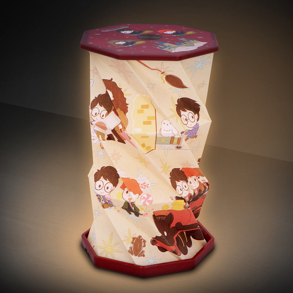 Paper lantern showing cartoon children with glasses reading, flying on a broom and riding a toy train, decorated with stars and sweets, lit from within and standing on a dark tabletop.