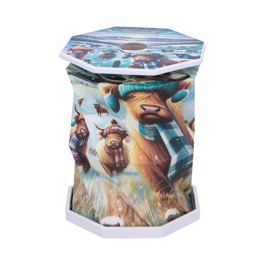Hexagonal collapsible lantern displays illustrated Highland cows wearing winter hats and scarves, set against snowy fields; white plastic top reads "POP GLOW" and hexagonal white base sits on white background.