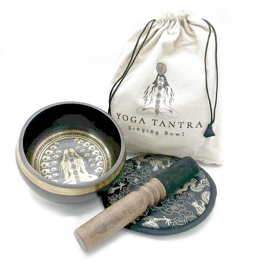 Singing bowl sits open revealing an etched meditating figure, resting beside a wooden mallet and embroidered cushion in front of a white drawstring bag labeled YOGA TANTRA Singing Bowl on a white background.