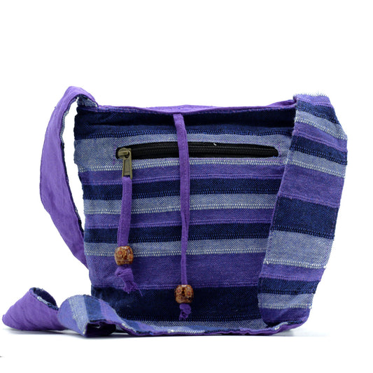 Handwoven purple-striped shoulder bag rests with its wide strap draped forward, showing a zip pocket and beaded drawstrings against a plain white background.